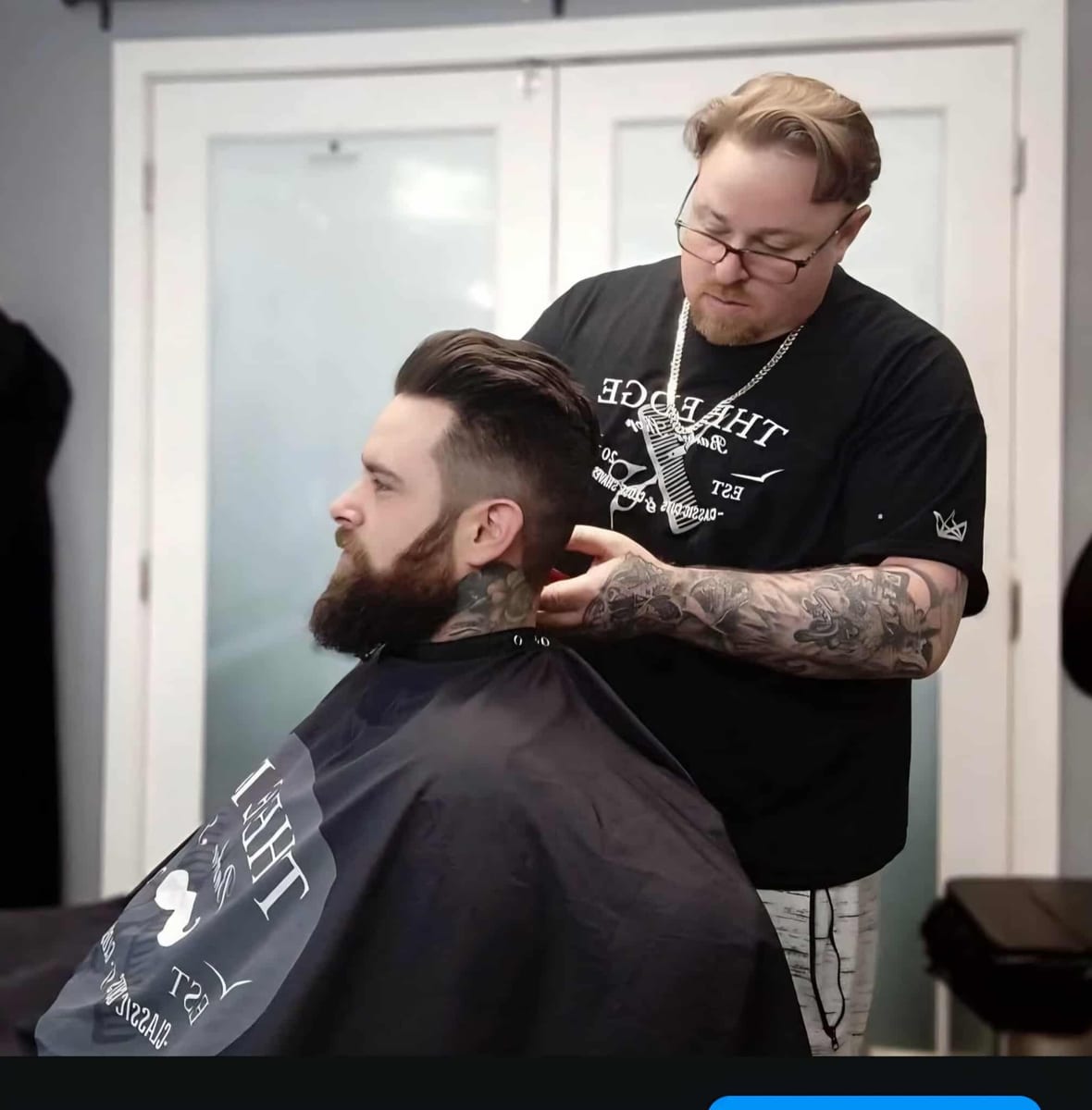 A barber in a THE EDGE shirt shaping a client's hair, the client's full beard and neck tattoo in profile